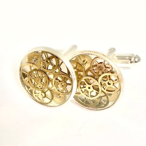 May include: Silver cufflinks with a clear, circular face. Each cufflink showcases an intricate design of small, gold-coloured gears, offering a mechanical, steampunk style. The cufflinks are presented against a white backdrop.