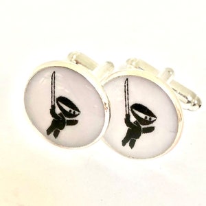 May include: A pair of silver cufflinks with a white background and a black silhouette of a ninja holding a sword.