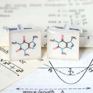 Puede incluir: Un par de gemelos de plata con fondo blanco y una fórmula química de cafeína impresa en negro, rojo y azul.