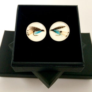 May include: A pair of cufflinks with a fly design. The cufflinks are silver and have a clear resin top with a fly encased inside. The fly is a brown and blue color with white feathers. The cufflinks are in a black box.