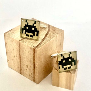 May include: A pair of gold cufflinks with a black and white pixelated alien design. The cufflinks are on a wooden surface.