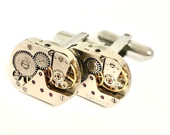 May include: A pair of silver cufflinks with exposed clockwork mechanisms. The cufflinks are round and have a silver finish. The clockwork is visible through the clear face of the cufflinks.