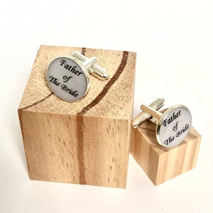 May include: A pair of silver cufflinks with a white background and black text that reads "Father of the Bride".