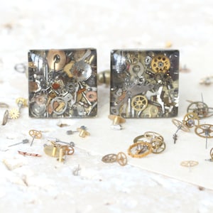 May include: A pair of square earrings with a clear resin base filled with assorted gold and silver watch parts. The earrings are shown on a white surface with loose watch parts scattered around them.