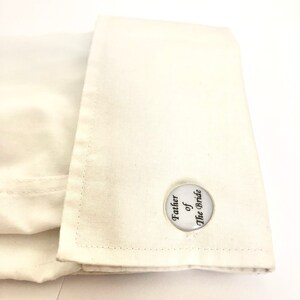 May include: White cufflinks with a round, silver-coloured design. The cufflinks feature the text "Father of the Bride" in black.