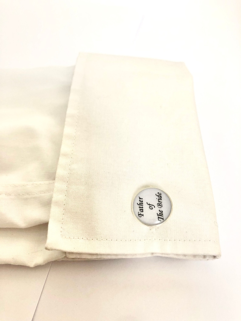 May include: White cufflinks with a round, silver-coloured design. The cufflinks feature the text "Father of the Bride" in black.