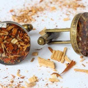 May include: A pair of cufflinks with a round, clear resin top filled with wood shavings. The cufflinks are made of brass and have a polished finish.