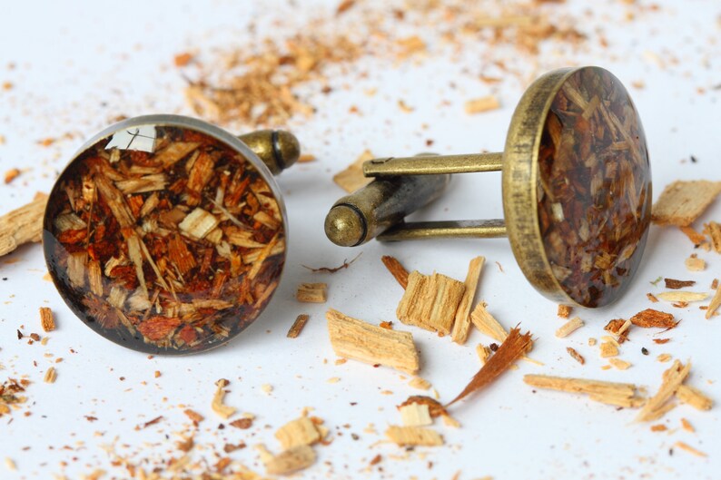 May include: A pair of cufflinks with a round, clear resin top filled with wood shavings. The cufflinks are made of brass and have a polished finish.
