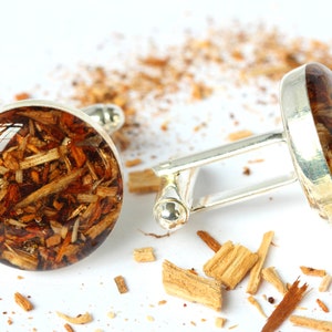 May include: A pair of silver cufflinks with round, clear resin insets containing wood shavings. The cufflinks are on a white surface with wood shavings scattered around them.