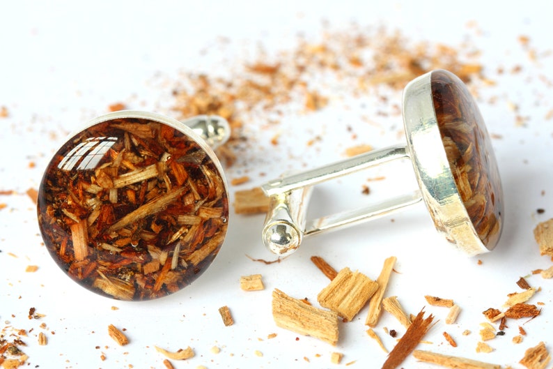May include: A pair of silver cufflinks with round, clear resin insets containing wood shavings. The cufflinks are on a white surface with wood shavings scattered around them.