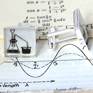 May include: A pair of silver cufflinks with a white background and a black and white illustration of a chemistry experiment. The experiment shows a flask with a tube leading to a beaker.