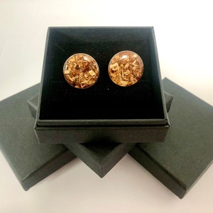 May include: A pair of round wood-filled resin earrings in a black gift box. The earrings are a light brown color with flecks of gold.