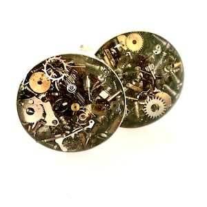 May include: A pair of round cufflinks with a steampunk design. The cufflinks are made of clear resin with embedded gears, screws, and other metal pieces.