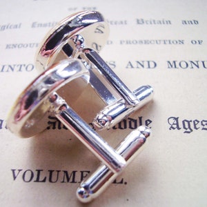 May include: A pair of silver cufflinks with a round, white, and yellow design. The cufflinks are on a white surface with text that reads "Volume I" and "Middle Ages".