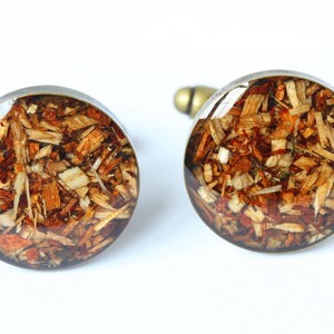 May include: A pair of round cufflinks with a silver metal base and a clear resin top. The resin contains a mixture of wood shavings in shades of brown, orange, and white.