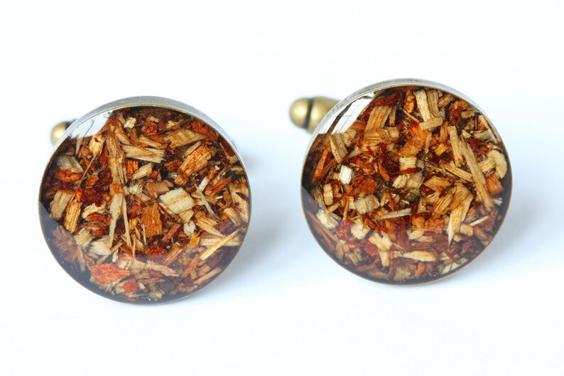 May include: A pair of round cufflinks with a silver metal base and a clear resin top. The resin contains a mixture of wood shavings in shades of brown, orange, and white.