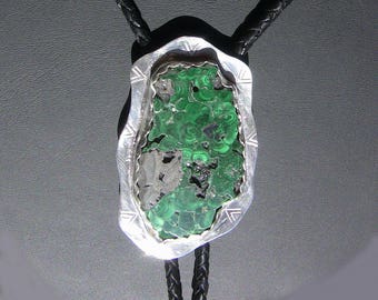 May include: A silver bolo tie with a green malachite stone set in a silver frame. The bolo tie has a black braided cord.