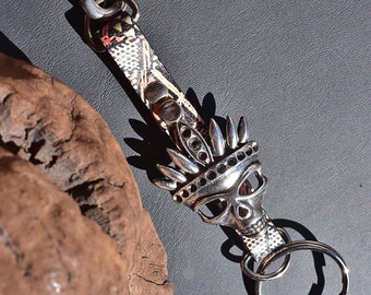 May include: A silver skull keychain with a Native American headdress design. The keychain is attached to a black and white patterned leather strap with a silver clasp.