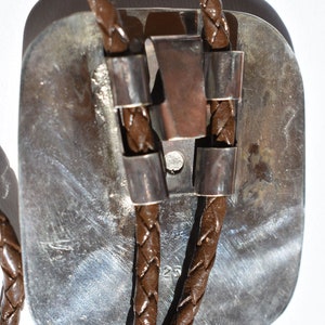May include: A bolo tie with a rectangular silver-toned pendant and brown braided leather cords. The pendant has a textured surface and is accented with silver-toned metal details. The cords are secured with silver-toned metal slides.