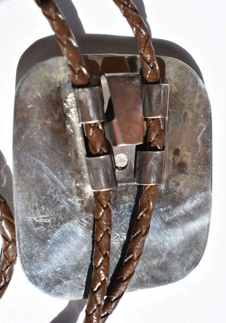 May include: A bolo tie with a rectangular silver-toned pendant and brown braided leather cords. The pendant has a textured surface and is accented with silver-toned metal details. The cords are secured with silver-toned metal slides.