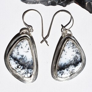 May include: A pair of silver dangle earrings. Each earring features a triangular, white stone with black and gray patterns, set within a silver frame. The edges of the stone have a serrated design. The earrings have a hook closure.