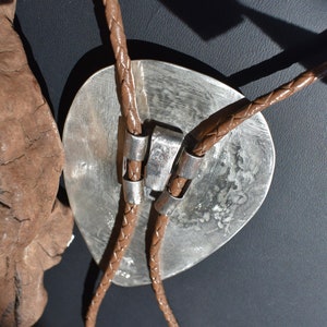 May include: A silver bolo tie with a large, textured oval pendant. The pendant is connected to a brown braided leather cord with silver accents. The bolo tie is a Western-style accessory.