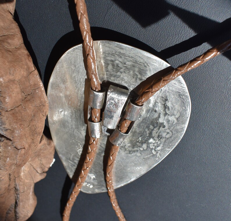 May include: A silver bolo tie with a large, textured oval pendant. The pendant is connected to a brown braided leather cord with silver accents. The bolo tie is a Western-style accessory.