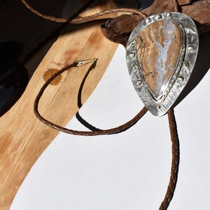 May include: A bolo tie with a teardrop-shaped pendant featuring a brown and white stone set in a silver frame. The braided leather cord is brown, and the tie is displayed on a wooden surface.
