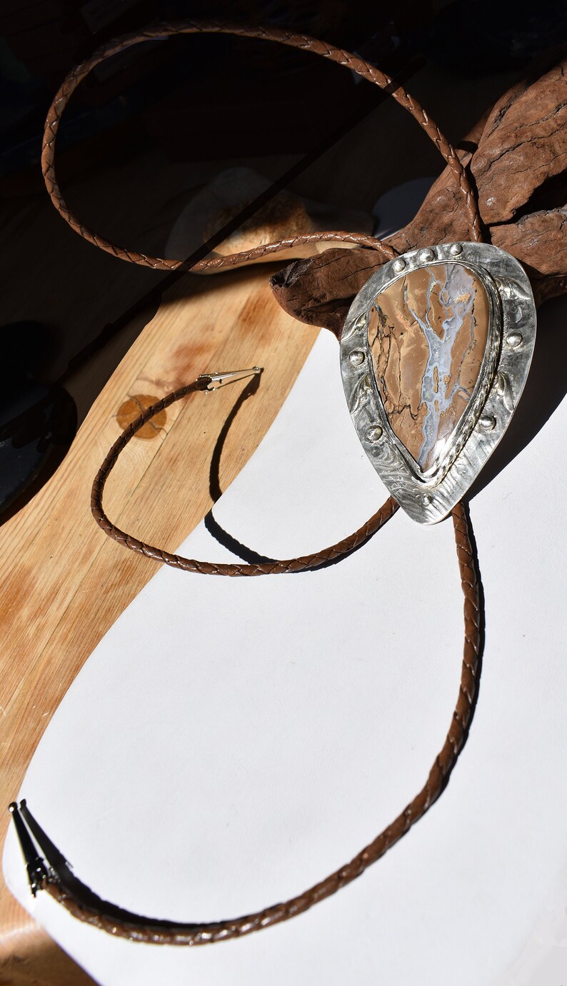 May include: A bolo tie with a teardrop-shaped pendant featuring a brown and white stone set in a silver frame. The braided leather cord is brown, and the tie is displayed on a wooden surface.