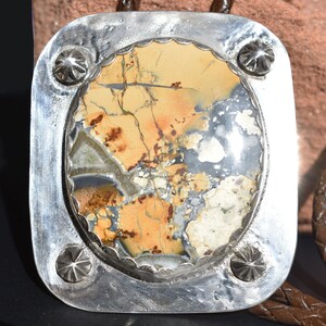 May include: A bolo tie with a rectangular silver pendant. The pendant features a polished, multi-colored stone with shades of tan, gray, and brown. The silver frame has a scalloped edge and decorative starburst accents.
