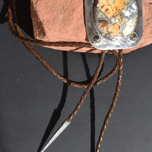 May include: A bolo tie with a rectangular silver pendant featuring a patterned stone in shades of beige, brown, and gray. The pendant is adorned with decorative studs. The braided brown leather cord has silver tips.