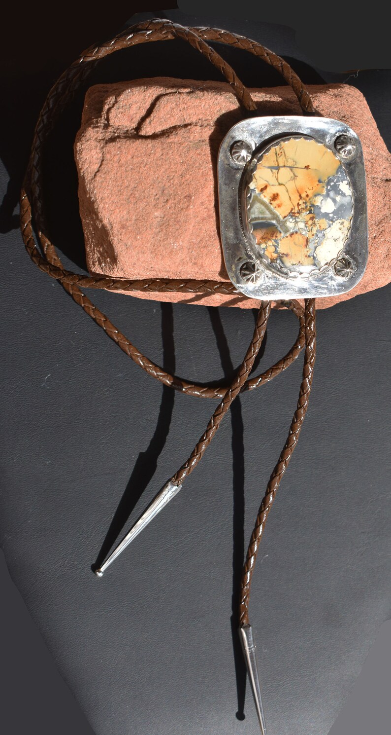 May include: A bolo tie with a rectangular silver pendant featuring a patterned stone in shades of beige, brown, and gray. The pendant is adorned with decorative studs. The braided brown leather cord has silver tips.