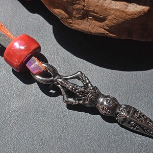 May include: A silver pendant with an ornate design, featuring a pointed end and a decorative handle. The pendant is attached to a leather cord with a red bead and colorful accents. The background is a dark gray surface with a piece of wood.