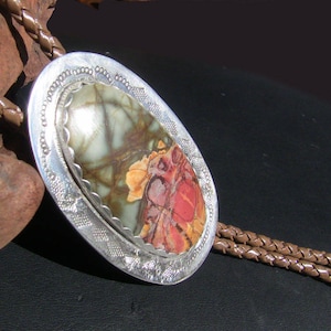 May include: A silver bolo tie with a brown leather cord and a large oval-shaped stone with a green, red, and white pattern.