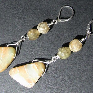 May include: A pair of dangle earrings featuring natural stone beads and a lever-back closure. The earrings have a silver-tone wire design, with a large, flat, light-colored stone at the bottom and three smaller, round, olive-green stones above.