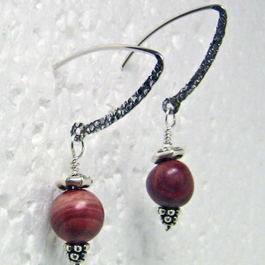 May include: A pair of handcrafted earrings featuring a silver hook and a textured design. Each earring has a round, reddish-brown bead with silver accents at the top and bottom. The earrings are displayed against a white background.