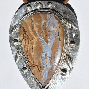 May include: A bolo tie with a large, teardrop-shaped stone pendant. The stone features a mix of brown, tan, and gray colors with a unique pattern. The pendant is framed by a silver metal border with decorative elements and a braided leather cord.