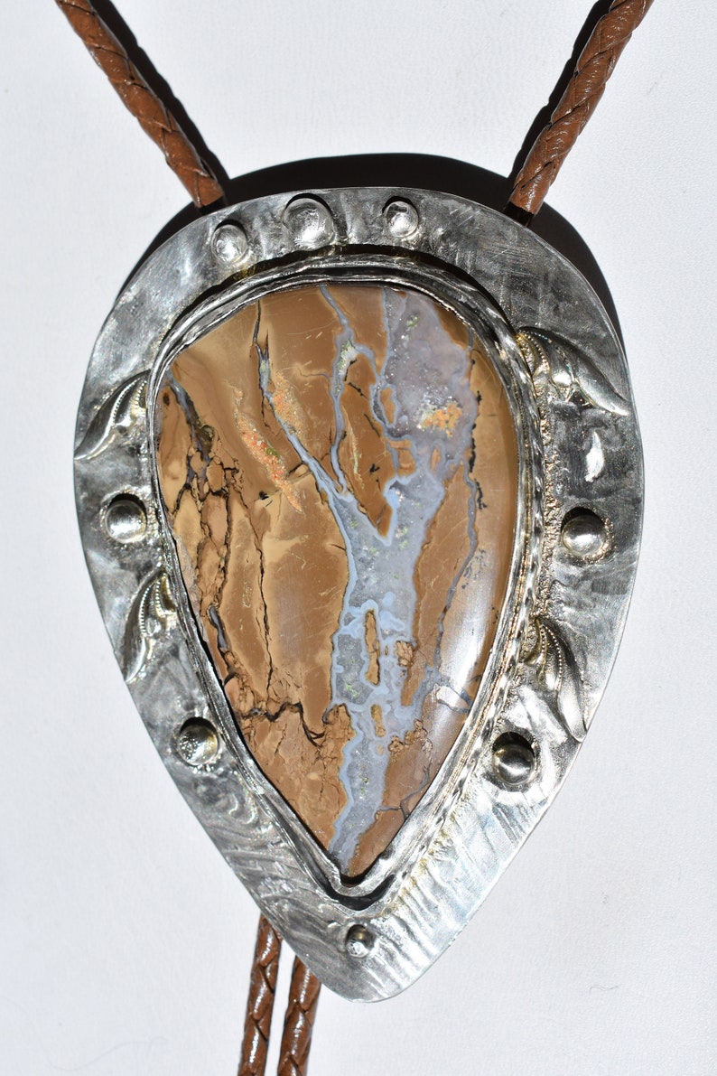 May include: A bolo tie with a large, teardrop-shaped stone pendant. The stone features a mix of brown, tan, and gray colors with a unique pattern. The pendant is framed by a silver metal border with decorative elements and a braided leather cord.
