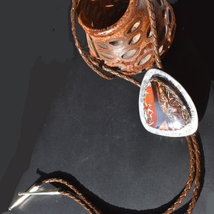May include: A brown leather bolo tie with a silver pendant featuring a colorful agate stone. The pendant is shaped like a teardrop and has a silver border.