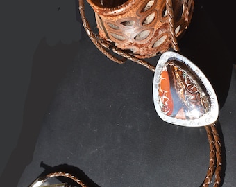 May include: A brown leather bolo tie with a silver pendant featuring a colorful agate stone. The pendant is shaped like a teardrop and has a silver border.