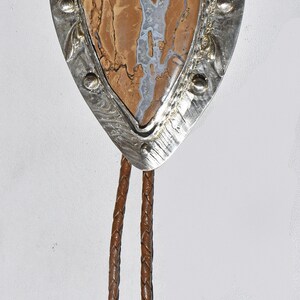May include: A bolo tie featuring a teardrop-shaped pendant with a brown and white stone set in a silver-toned metal frame. The braided leather cord is brown.