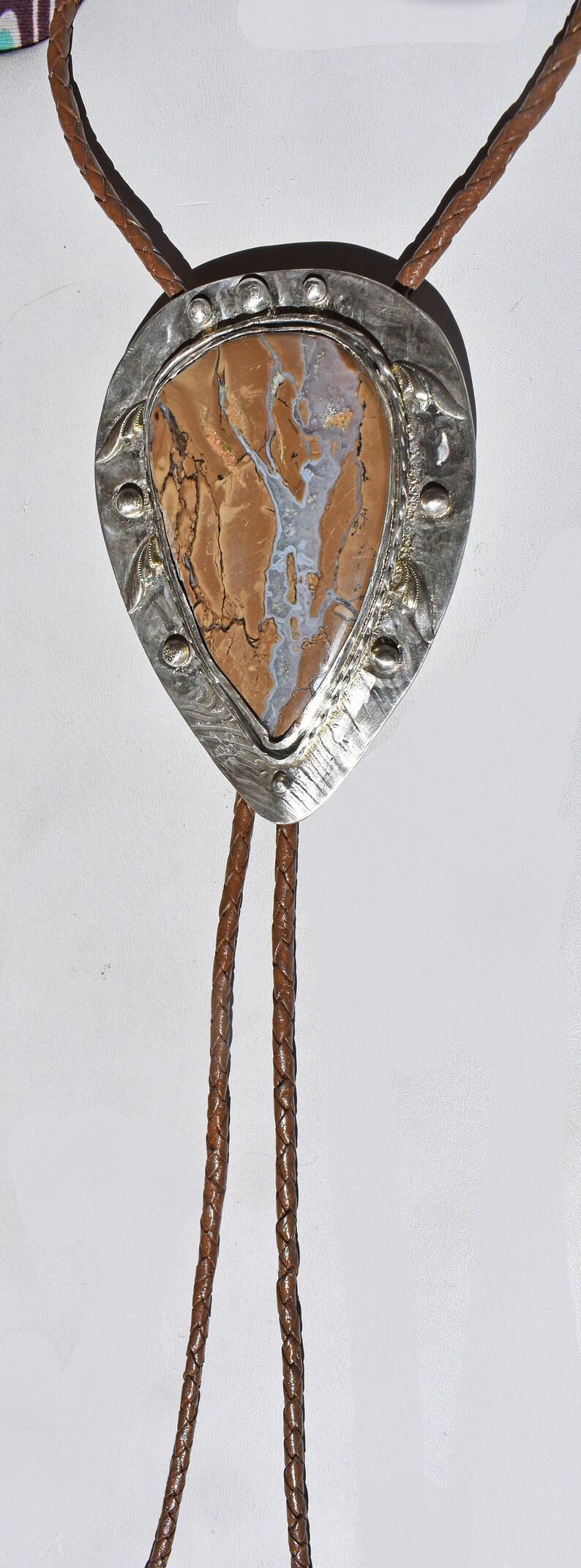May include: A bolo tie featuring a teardrop-shaped pendant with a brown and white stone set in a silver-toned metal frame. The braided leather cord is brown.