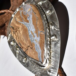 May include: A bolo tie with a silver-toned metal setting. The centerpiece is a brown and white stone with a unique pattern. The braided leather cord is brown. The bolo tie is displayed against a white background and a piece of driftwood.