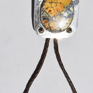 May include: A bolo tie featuring a rectangular silver pendant with a natural stone inlay in shades of yellow, brown, and gray. The pendant is attached to a braided brown leather cord with silver tips. A stylish accessory.