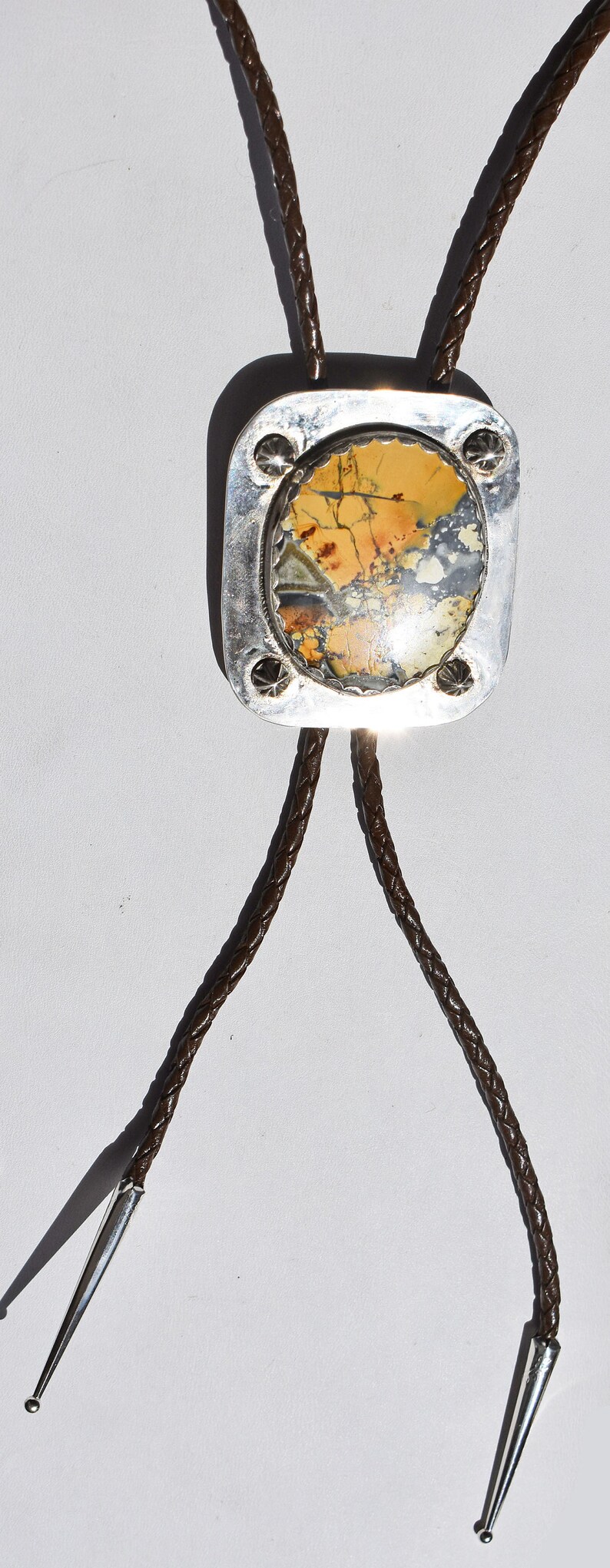 May include: A bolo tie featuring a rectangular silver pendant with a natural stone inlay in shades of yellow, brown, and gray. The pendant is attached to a braided brown leather cord with silver tips. A stylish accessory.