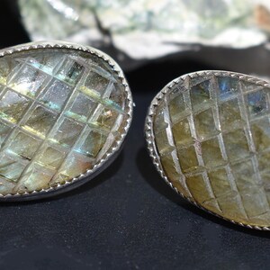 May include: A pair of oval-shaped earrings with a silver border. The earrings feature a grid-like pattern carved into a light green and yellow stone. The edges of the silver border have a scalloped design.