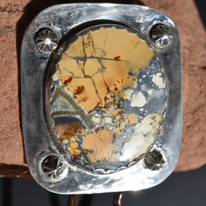 May include: A silver bolo tie with a rectangular, patterned stone center. The stone features shades of tan, brown, and gray. The silver frame has a decorative edge and star-shaped accents. A brown leather cord completes the design.