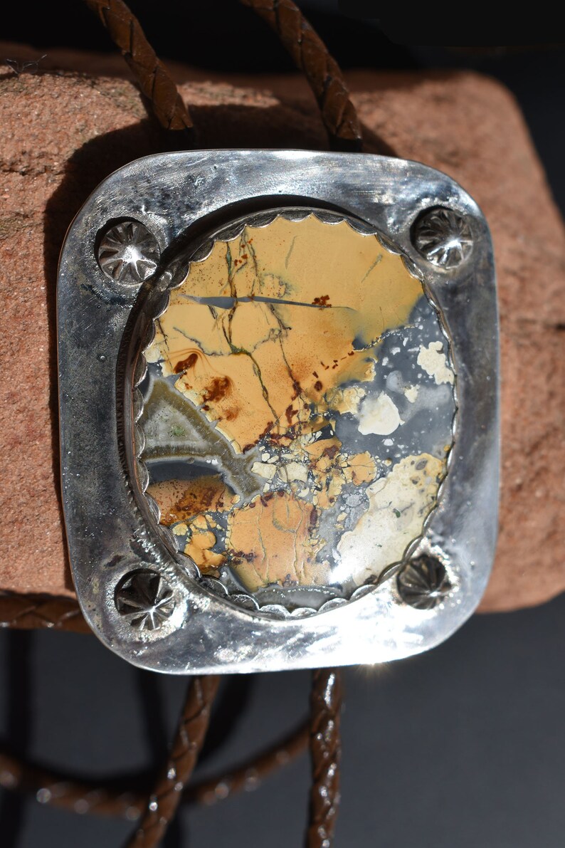 May include: A silver bolo tie with a rectangular, patterned stone center. The stone features shades of tan, brown, and gray. The silver frame has a decorative edge and star-shaped accents. A brown leather cord completes the design.