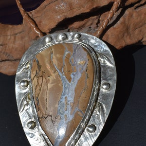 May include: A bolo tie featuring a large, teardrop-shaped stone with brown, beige, and white patterns, set in a silver-colored metal frame. The frame has a decorative edge and is attached to a braided brown leather cord.