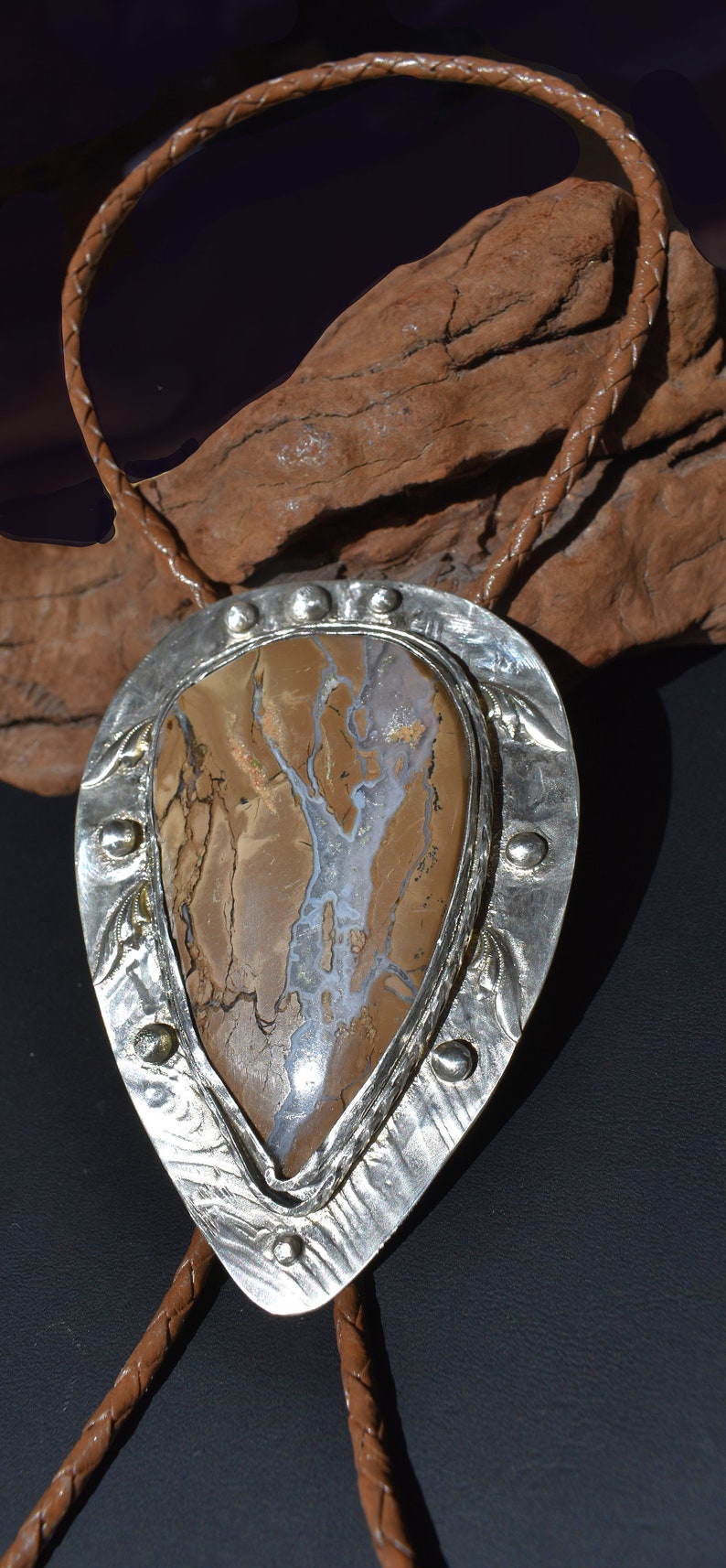 May include: A bolo tie featuring a large, teardrop-shaped stone with brown, beige, and white patterns, set in a silver-colored metal frame. The frame has a decorative edge and is attached to a braided brown leather cord.